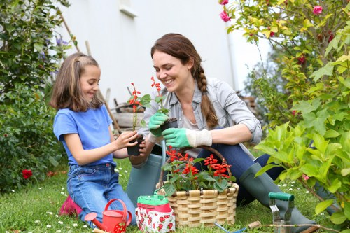 A woman and a young girl are gardening together in a well-maintained backyard, with lush green grass and various flowering plants and shrubs. The woman, dressed in casual outdoor clothes and green gardening gloves, is smiling as she helps the girl plant a small potted plant with red flowers. The girl, wearing a blue t-shirt and denim shorts, is engaged in the activity, holding the pot. Nearby, a woven basket filled with gardening tools and supplies is visible, along with a small watering can. The garden features a mix of vibrant flowers, leafy plants, and a backdrop of trees, with sunlight illuminating the scene, indicating a bright, clear day. This outdoor space showcases thoughtful planting and landscaping, typical of residential gardens in Southwark, suitable for nurturing gardening skills and maintaining a healthy, attractive outdoor environment as part of professional gardening services offered by Landscapers Southwark.