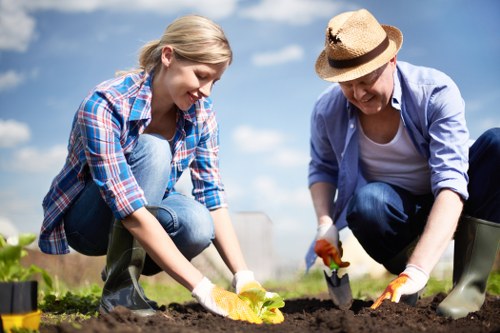 A young woman and an older woman are working together outdoors in a garden, planting seedlings in prepared soil. The young woman, dressed in a blue plaid shirt and jeans, is kneeling and carefully placing a small plant into the ground, while the older woman, wearing a light blue jacket, white top, and protective hat, is also kneeling and using a trowel to assist with the planting. They are both wearing gardening gloves and are surrounded by garden beds with dark, rich soil. In the background, there is a clear blue sky with a few clouds, and the garden appears well-maintained with some leafy green plants nearby. This scene takes place in a sunny outdoor environment that suggests careful gardening and planting activities, typical of a landscaped backyard or community garden in Southwark, enhancing sustainable outdoor space development by Landscapers Southwark.
