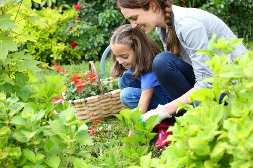 A woman and a young girl are gardening together in a lush outdoor garden setting; they are crouched down near a variety of green plants, including leafy bushes and possibly flowering shrubs. The woman, with long brown hair in pigtails, is wearing a light grey top and blue jeans, while the girl is dressed in a blue t-shirt and denim shorts. They are smiling and focused on the plants, with the girl holding a woven basket, suggesting they are harvesting or collecting garden produce. The garden features a well-maintained lawn area with dense foliage, vibrant green leaves, and red flowering plants, all situated in a natural outdoor environment that suggests late spring or summer weather conditions. The scene exudes a peaceful, productive gardening activity, indicative of professional outdoor maintenance and gardening services like those offered by Landscapers Southwark, supporting healthy, attractive garden spaces in Southwark and surrounding areas near the postcode area SE.