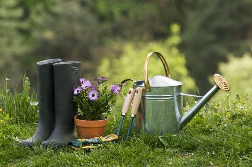 A pair of black rubber gardening boots standing upright on a lush green lawn, accompanied by a small terracotta pot filled with purple flowering plants, gardening gloves, and a trowel. Behind these items, there is a silver metal watering can with a brass handle and a long spout, positioned on the grass. The background features a natural outdoor setting with blurred green foliage and trees, indicating a well-maintained backyard garden or outdoor space. The scene suggests preparation for gardening activities common in residential gardens within Southwark, under natural daylight with clear weather, highlighting outdoor maintenance elements associated with lawn and garden care services.