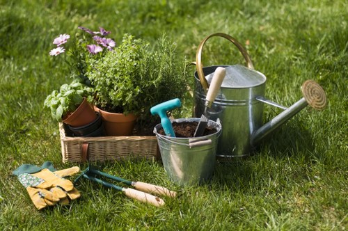 A neatly maintained backyard garden with a lush green lawn, bordered by a variety of potted plants including purple and green foliage. In the foreground, gardening tools such as a pair of yellow work gloves, a trowel, and a small fork rest on the grass. Adjacent to the tools, there is a shiny metal watering can with a wooden handle, and a small metal bucket containing a blue-handled trowel and other gardening implements. Behind these, a rectangular wicker basket holds several potted plants, including flowering varieties with purple blooms, and green leafy shrubs. The overall scene is outdoors, captured on a bright, slightly overcast day, with natural daylight illuminating the garden, which suggests a well-cared-for space suitable for gardening and outdoor maintenance activities common to Southwark households. This image evokes the type of gardening and landscaping services typical of Southwark, including plant care, lawn maintenance, and outdoor tool management, as part of professional garden upkeep by Landscapers Southwark.