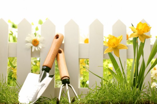 A close-up outdoor scene of a well-maintained garden area in front of a white picket fence, featuring a lush green lawn with dense, healthy grass, alongside blooming yellow daffodils and white daisies growing at the edge of the garden bed. In the foreground, two gardening tools— a hand trowel and a small hand rake with wooden handles—rest on the grass, suggesting recent or upcoming gardening activity. The background reveals soft natural light filtering through the fence, indicating a bright, clear day with a garden space that is cared for and inviting. This setting exemplifies typical features of residential gardens in Southwark, making it relevant for landscaping, lawn care, and gardening services offered by Landscapers Southwark, with a focus on outdoor maintenance and plant care in the local area.