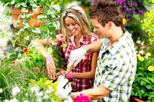 The image shows a brightly lit outdoor garden space with a lush, densely planted flower bed surrounded by green foliage, including various flowering plants with vibrant pink, purple, and yellow blossoms. In the foreground, a well-maintained grassy lawn with a uniform, dense appearance extends across the scene. The background features a hedge or shrub border that separates the garden area from neighboring greenery, and there are some potted plants hanging from wooden trellising structures. The setting appears to be part of a residential backyard, likely in the Southwark area, given the context. The garden surface includes a combination of grass, soil, and paved pathways made of stone or concrete, providing clear walking routes through the space. The natural lighting suggests a bright, sunny day with minimal cloud cover, contributing to the vibrant colours and shadows cast across the garden. This scene exemplifies typical garden maintenance work that a landscaping and gardening service like Landscapers Southwark might provide, including plant care, lawn upkeep, and outdoor beautification to enhance the outdoor environment of a suburban property.