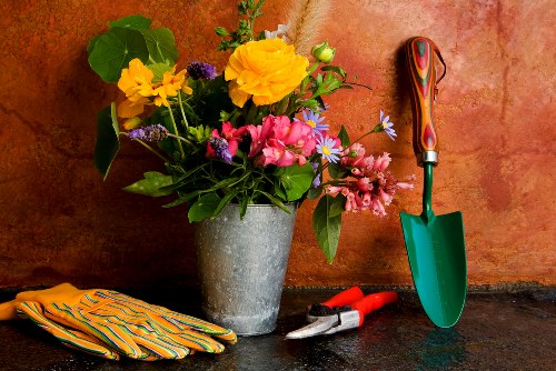 This image displays a collection of gardening tools and decorative items arranged on a dark surface against a warm-toned, textured wall. Prominently, there is a galvanized metal bucket filled with a variety of vibrant flowers, including yellow, pink, purple, and white blossoms, surrounded by lush green leaves. To the right of the flower arrangement, a small green trowel with a wooden handle is positioned upright, its blade resting on the surface. Adjacent to the trowel, there are a pair of pruning shears with red handles and a garden rake with a wooden handle, both placed in a resting position. On the left side, a pair of striped gardening gloves are neatly folded, featuring bright yellow, green, and orange hues. The scene evokes a professional gardening and landscaping environment, with a subtle connection to outdoor maintenance services offered by companies like Landscapers Southwark, focusing on garden decoration, planting, and general lawn care activities within the Southwark area in London.