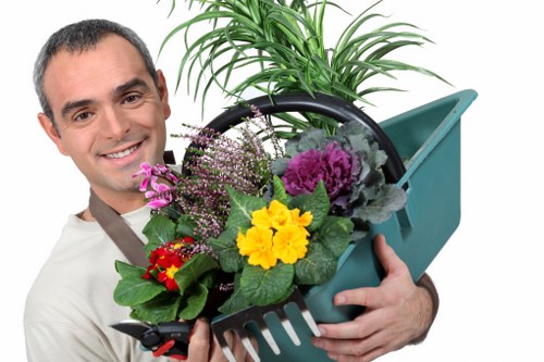 A man with a friendly expression is holding a blue gardening trowel filled with a variety of colorful flowering plants and green foliage, including purple, yellow, red, and pink blooms, in an outdoor garden setting. Behind him, there is a lush, green potted plant with long, arching leaves, placed on a paved or concreted surface that appears well-maintained. The garden environment suggests a neat and cared-for outdoor space, likely part of a residential garden in Southwark, featuring a mix of flowering plants, shrubs, and a healthy lawn area visible in the background. The scene reflects outdoor gardening activities, emphasizing plant care and landscape maintenance typical of a professional gardening service, with natural sunlight enhancing the vibrant plant colors and greenery.