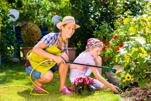 In a lush, well-maintained garden with vibrant greenery and flowering plants, a woman and a young girl are engaged in outdoor gardening activities. The woman, wearing a straw hat, yellow apron, and casual clothing, is smiling as she holds a garden hose, watering the flower beds. The girl, dressed in a white top, pink raincoat, and a patterned hat, is focused on planting or tending to the colorful flowers, which include pink and yellow blooms. The garden features a neatly trimmed lawn with lush grass, bordered by flower beds filled with various flowering plants and shrubs. In the background, there are decorative garden lights and additional foliage, creating a pleasant, inviting atmosphere. The scene suggests a warm, sunny day, ideal for outdoor gardening, reflecting professional gardening and landscaping services available in Southwark, with natural textures of soil, grass, and plant foliage enhancing the visual appeal of the landscaped outdoor space for residential or community gardening projects.
