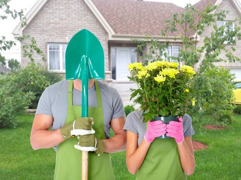 In a suburban front garden, a person wearing a teal garden shovel helmet, a grey t-shirt, and an olive green apron is standing on a well-maintained lawn with neatly trimmed grass, holding a large green garden spade. Next to them, another individual in a grey t-shirt and bright pink gardening gloves is holding a vibrant yellow-flowering shrub with lush green foliage, partly covering their face. The garden features a balanced layout with a green lawn in the foreground, bordered by a flower bed with dark mulch and ornamental plants. In the background, a light grey brick house with white-framed windows and a bay window is visible, complemented by a small hedge and young tree with light green leaves. The scene is outdoors on a clear day with natural lighting, and the setting suggests a focus on gardening and outdoor maintenance activities, consistent with landscaping services such as patio repairs or garden improvements offered by Landscapers Southwark.