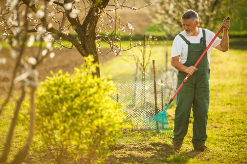 A man dressed in gardening overalls is raking soil in a landscaped garden area in Southwark, with a large flowering tree and a neatly maintained flower bed visible in the background. The garden features a lush green lawn, bordered by a wire fence separating the flower bed from the rest of the yard. The scene is illuminated by natural sunlight, suggesting a bright, clear day, with some blossoming branches on the tree indicating springtime. The man appears focused on outdoor maintenance, supporting gardening and landscaping services provided by landscapers southwark.co.uk, as part of a professional outdoor care arrangement in a typical suburban garden setting. The garden's layout emphasizes a combination of cultivated beds and open grassy areas, with different textures from mulch to soil, and natural plant colours ranging from vibrant greens to soft pinks in the blossoms.