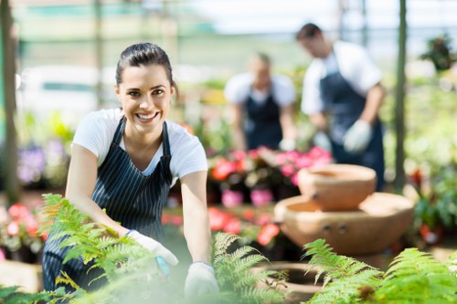 A young woman with a bright smile, wearing a striped apron, is tending to a lush garden bed within a greenhouse or conservatory setting in Southwark. She is surrounded by green, healthy ferns and other leafy plants, with her hands on the soil, indicating active gardening or planting work. The background features blurred images of two other gardeners, a man and a woman, wearing aprons and working near vibrant flower displays and stacks of terracotta pots. The environment is illuminated by natural sunlight streaming through large glass panes, creating a warm and inviting atmosphere typical of professional gardening environments and outdoor maintenance sites. The scene reflects a professional landscaping or gardening service focus, emphasizing attention to detail and plant care, suitable for SEO related to lawn and garden maintenance services in Southwark or the surrounding London area.